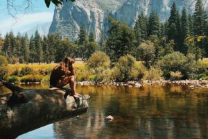 Woman in the wilderness sitting on a log over a river with tall mountain backdrop